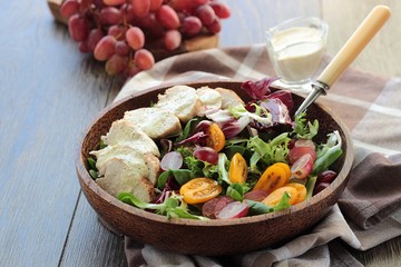 Grilled chicken fillet, pink grapes and tomatoes with colorful salad mix with lettuce leaves, chicory, corn salad and radicchio in a wooden bowl on a table.