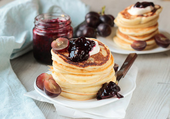 Stack of homemade pancakes on a white plate with sour cream, jam and grapes.