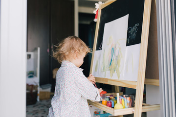 Portrait of adorable baby girl drawing with paints at easel .