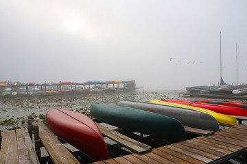 Morning autumn nature misty scene. Scenic view with rising sun penetrate through deep morning fog over the lake, wooden pier  and boats on a foreground, rack with kayaks on a background. 