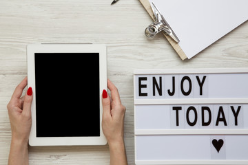 Fototapeta na wymiar Work space with tablet, notepad and 'Enjoy today' word on lightbox over white wooden background, top view. Female hands hold tablet with blank screen. From above, flat-lay, overhead.