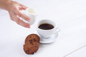 A woman's hand pours milk into coffee. A cup of fragrant coffee and cookies on a white background.
