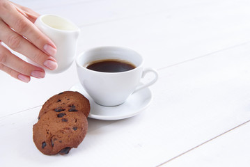 A woman's hand pours milk into coffee. A cup of fragrant coffee and cookies on a white background.