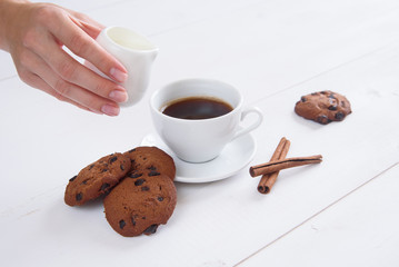 A woman's hand pours milk into coffee. A cup of fragrant coffee with cinnamon and cookies on a white background.