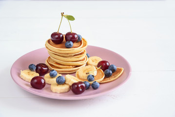 Tasty homemade pancakes with fresh berries on violet plate on a white background. Healthy breakfast of pancakes with blueberries, cherries and bananas.