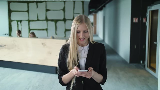 Young formal woman using smartphone in office. Stylish blond woman in jacket standing in contemporary office hall and surfing smartphone with smile.