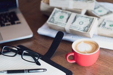 Close up of coffee cup, glasses, money, notepad, pen and laptop on wooden table