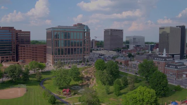 drone flying over Shaw Park in Clayton with swimming pool and baseball diamond. Camera pulls back along city buildings, clouds and blue sky.