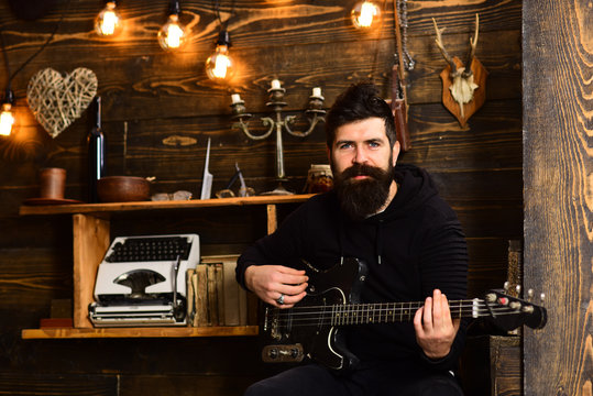 Man bearded musician enjoy evening with bass guitar, wooden background. Soul music. Man with beard holds black electric guitar. Guy in cozy warm atmosphere play relaxing soul music