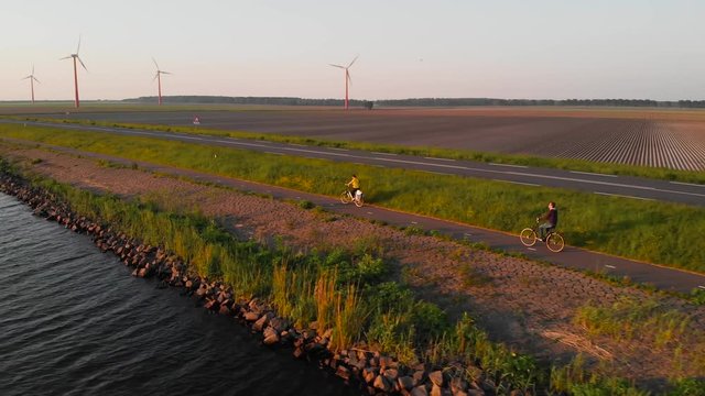 Couple enjoying a bicycle ride on a bike path along shore of Dutch polder Flevoland, with windmill park and fields on the background, at sunset.