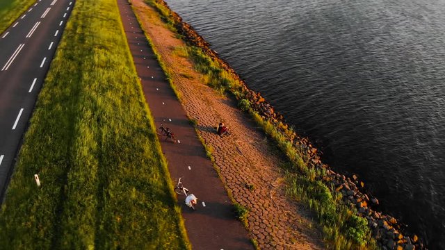 Aerial shot of a romantic couple relaxing and sitting on a chore of Dutch polder Flevoland watching sunset, with their bicycles standing behind on a bike path and road on the background