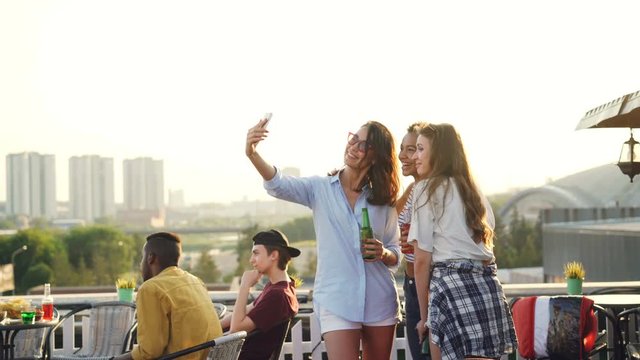 Cheerful female friends are taking selfie with smartphone and laughing standing on rooftop with drinks in bottles enjoying party. Technology, beverage and youth concept.