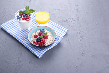 Delicious and healthy breakfast, gray background and napkin. Yogurt, muesli, granola, raspberry and blueberry berries and fresh orange juice. Copy space.