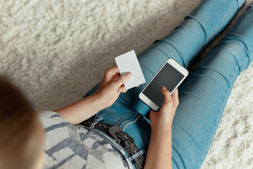 Young woman holding credit card and using phone sitting on floor. Payment.  Online shopping concept.Concept of blogging. Modern technologies Top view.