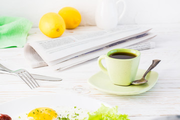 A cup of black coffee, a newspaper and cutlery on a white wooden table served for breakfast