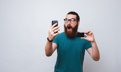 Studio shot of shocked and terrified young european man holding credit card and smartphone, standing with opened mouth from amazement over white background. Someone stole money from my account