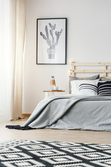 Patterned carpet and poster in grey simple bedroom interior with blanket on bed. Real photo