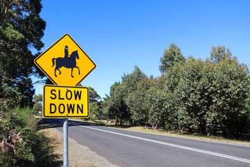 a black and yellow Slow Down, Horse Riders warning sign near a stretch of road