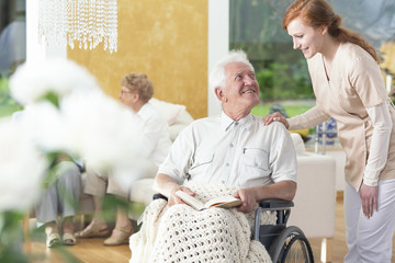 A man in his old age ia a wheelchair talking to a personal assistant in a common room of a rehabilitation center.