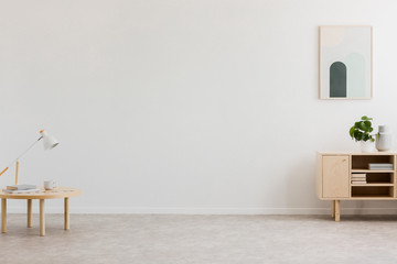 Desk lamp on a small table and a simple, wooden cabinet in an empty living room interior with white wall and place for a sofa. Real photo.