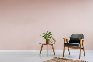 Wooden armchair with gray upholstery and a side table in a pastel pink living room interior with place for a bookcase. Real photo.