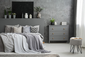 Monochromatic grey bedroom interior with a big bed with throws and pillows and a drawer cabinet...