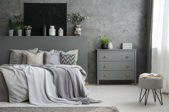Monochromatic grey bedroom interior with a big bed with throws and pillows and a drawer cabinet against a wall with a black canvas. Footstool with an open book. Real photo