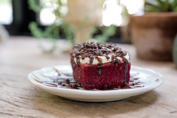 A piece of red velvet cake with cream and chocolate in white ceramic plate on wooden table