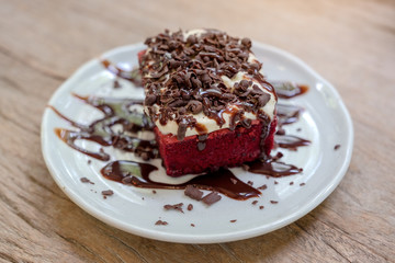 A piece of red velvet cake with cream and chocolate in white ceramic plate on wooden table
