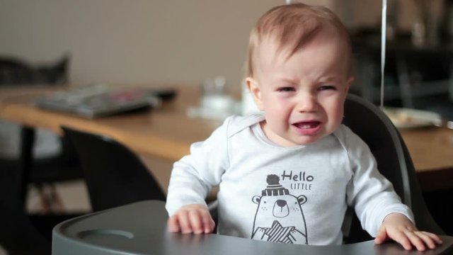 Little crying baby sitting on high chair indoor
