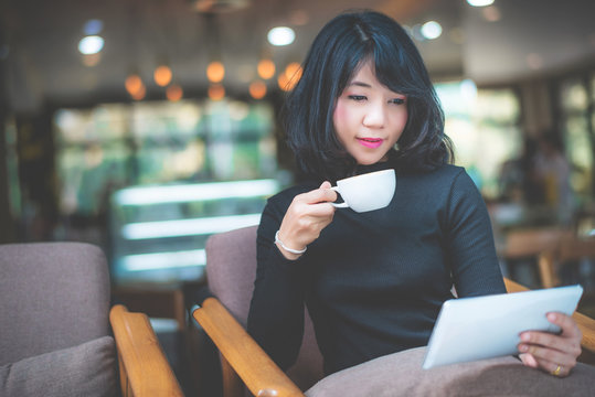 Beautiful attractive young Asian woman holding a cup of coffee in hand and sitting on sofa at cafe in the morning, vintage color tone. 