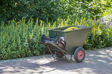 grosse grüne Schubkarre mit Wasserschlauch und Werkzeug auf Weg vor Blumenbeet