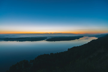 View from high shore on broad river. Riverbank with forest under thick fog. Dawn reflected in water. Yellow glow in picturesque predawn sky. Colorful morning atmospheric landscape of majestic nature.