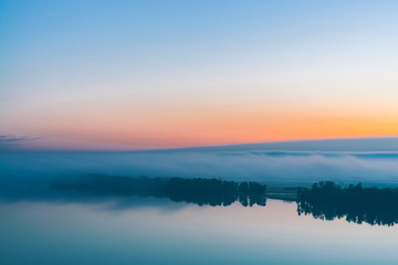 Fototapeta na wymiar Broad mystical river flows along diagonal shore with silhouette of trees and thick fog. Orange and pink glow in predawn vanila sky. Morning atmospheric landscape of majestic nature in tender tones.