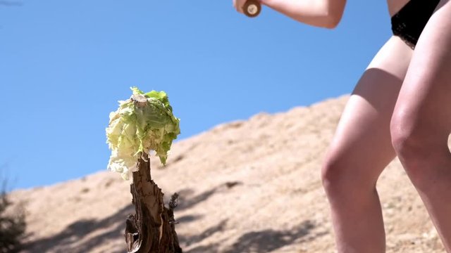 Slow motion: Sexy young girl in a black swimsuit and glasses, destroys fruits and vegetables with a baseball bat. It is located on a sandy beach near the sea in summer. It breaks cabbage