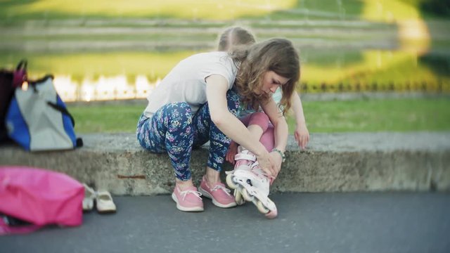 Mother helps his daughter to wear a helmet and protective gear, for roller skating in the park. Woman helps girl put on protective knee and elbow pads. Active family rest in the park.