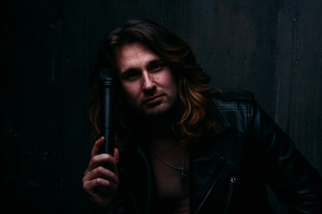 brutal man in a leather jacket on a naked body with long hair. a lonely man is sad and desperate. holds a microphone in his hand, leans against the wall