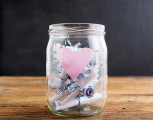 Einmachglas mit rosa Herz und zusammengerollten Notizzetteln auf einem Holztisch, schwarzer...