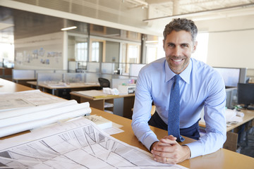 Male architect leaning on desk with plans looking to camera