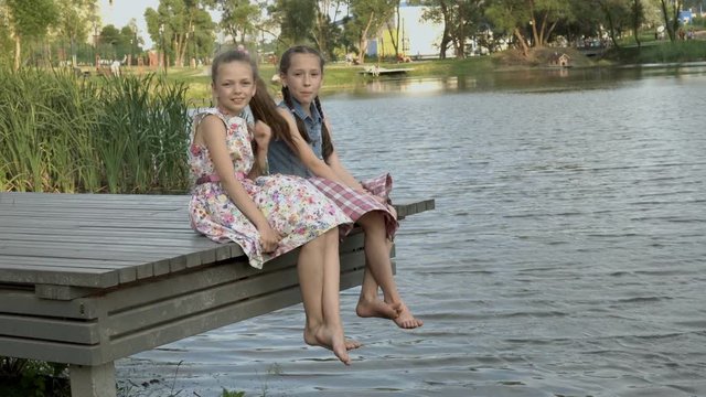 Two happy little girls are sitting in the summer on a wooden bridge by the river in the city Park next to each other with their bare feet over the water. They look at the camera and laugh.