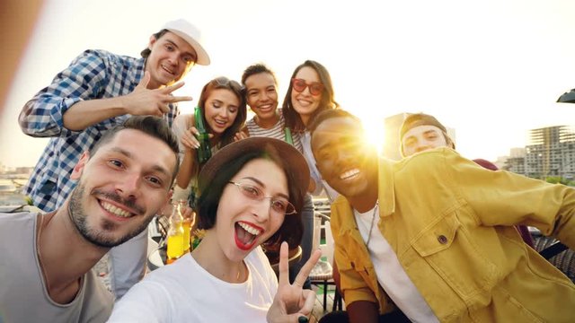 Point of view shot of pretty lady in trendy hat holding camera and taking selfie with friends enjoying rooftop party with soft drinks, people are looking at camera and laughing.