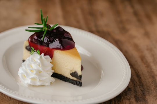 Delicious and sweet blueberry New York cheesecake on white plate served with whipped cream on wood table in close up view with copy space. Homemade bakery for cafe and restaurant or birthday cake.