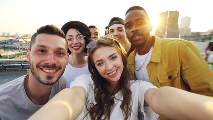 Attractive young woman is taking selfie with friends on rooftop, girl is holding camera and posing while her mates are having fun making funny faces and gestures. - Powered by Adobe