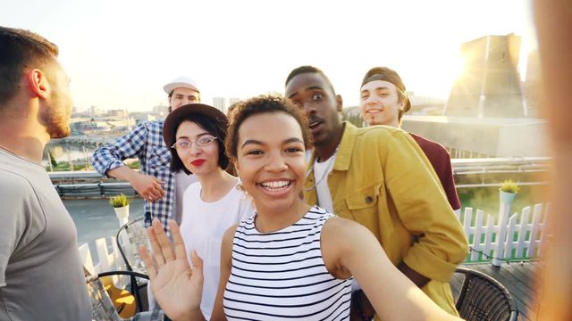 Cheerful African American girl is taking selfie adjusting camera then calling her friends, posing and laughing at rooftop party. Modern city is visible in background.