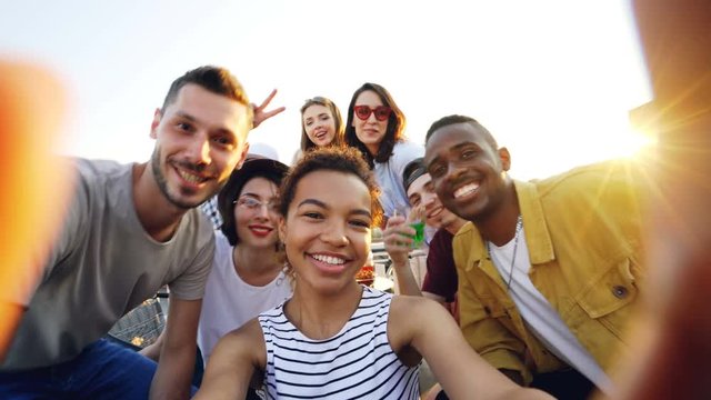 Point of view shot of African American girl holding camera and taking selfie with happy friends at party on roof. Men and women are looking at camera, posing and laughing.
