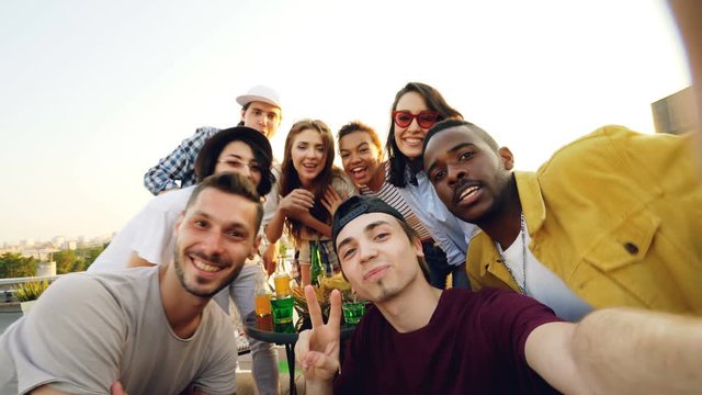 Point of view shot of joyful young man adjusting camera then calling his friends to take selfie with drinks at roof party, people are laughing and posing on sunny day.