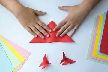 Детские руки делают бабочку оригами из красного листика бумаги на белом фоне. Обучение детей