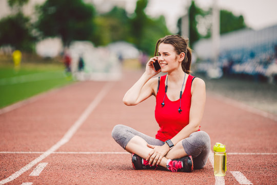 Sport and technology. A beautiful young Caucasian woman with ponytail sitting resting after workout during run at stadium, a red treadmill track. Uses for cell phone headphones and sports watches