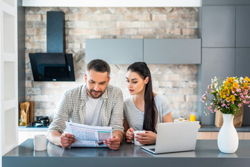 portrait of married couple reading newspaper together at counter with laptop in kitchen