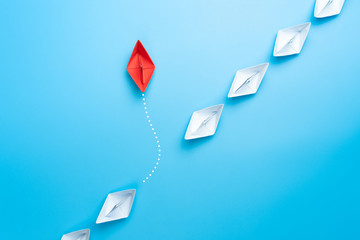 Group of white paper ship in one direction and one red paper ship pointing in different way on blue...